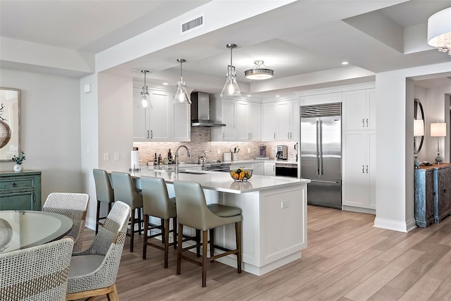 kitchen with white cabinets, kitchen peninsula, wall chimney exhaust hood, decorative light fixtures, and stainless steel built in refrigerator