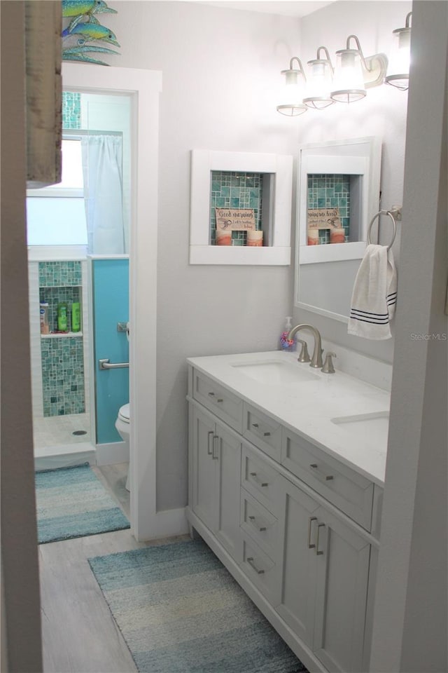 bathroom featuring double vanity, toilet, a sink, wood finished floors, and tiled shower