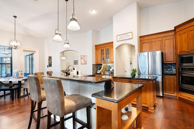 kitchen with a kitchen island with sink, appliances with stainless steel finishes, pendant lighting, dark wood-type flooring, and decorative backsplash