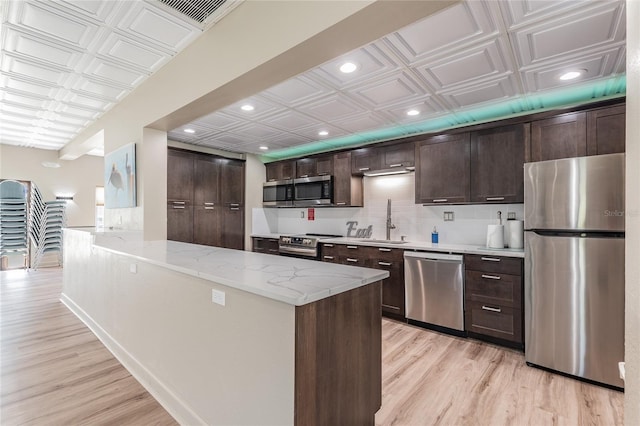 kitchen with light stone countertops, appliances with stainless steel finishes, dark brown cabinetry, sink, and light hardwood / wood-style floors