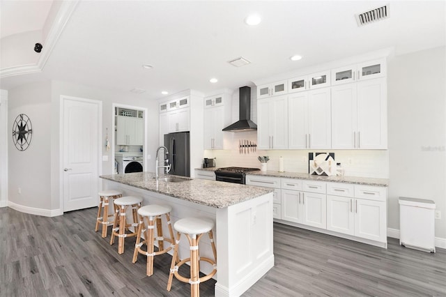 kitchen with black gas range, wall chimney range hood, sink, an island with sink, and white cabinetry