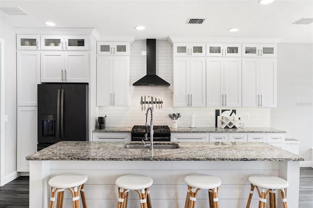 kitchen with white cabinets, stainless steel fridge, an island with sink, and wall chimney range hood