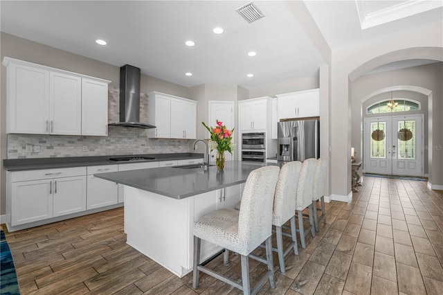 kitchen with white cabinetry, french doors, wall chimney exhaust hood, stainless steel appliances, and an island with sink