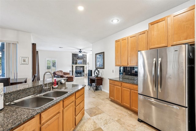 kitchen featuring stainless steel refrigerator, ceiling fan, sink, dark stone countertops, and a fireplace