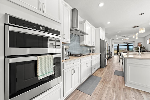 kitchen featuring white cabinetry, stainless steel appliances, decorative backsplash, hanging light fixtures, and wall chimney range hood