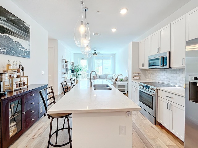 kitchen featuring a center island with sink, sink, appliances with stainless steel finishes, decorative light fixtures, and white cabinetry