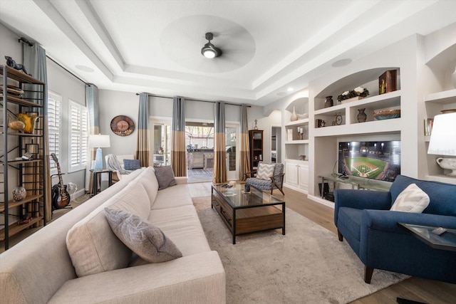 living room featuring ceiling fan, a raised ceiling, built in features, and light wood-type flooring