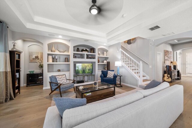 living room with a raised ceiling, ceiling fan, built in features, and light wood-type flooring