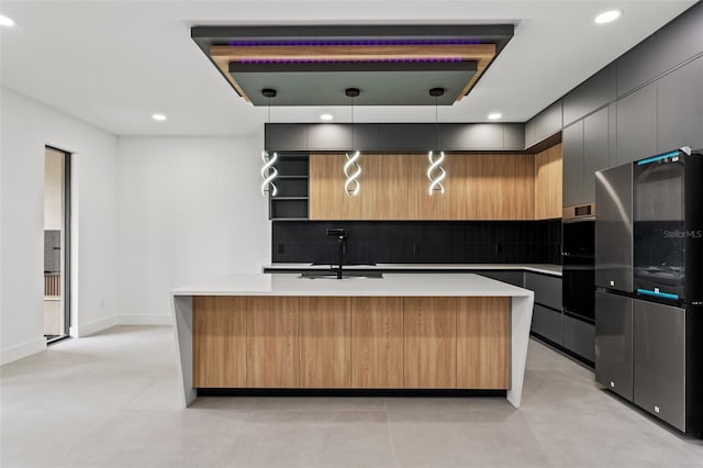 kitchen featuring sink, an island with sink, double oven, decorative light fixtures, and stainless steel refrigerator