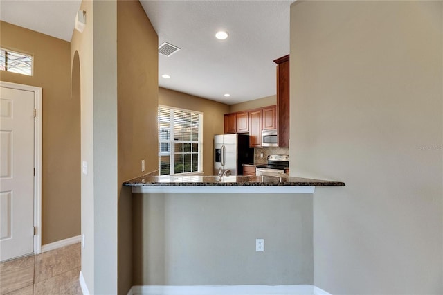 kitchen with kitchen peninsula, dark stone countertops, decorative backsplash, light tile patterned floors, and appliances with stainless steel finishes