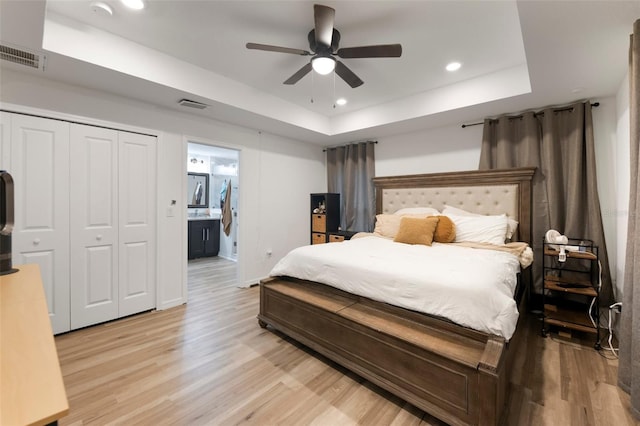 bedroom featuring ensuite bathroom, ceiling fan, a tray ceiling, light wood-type flooring, and a closet