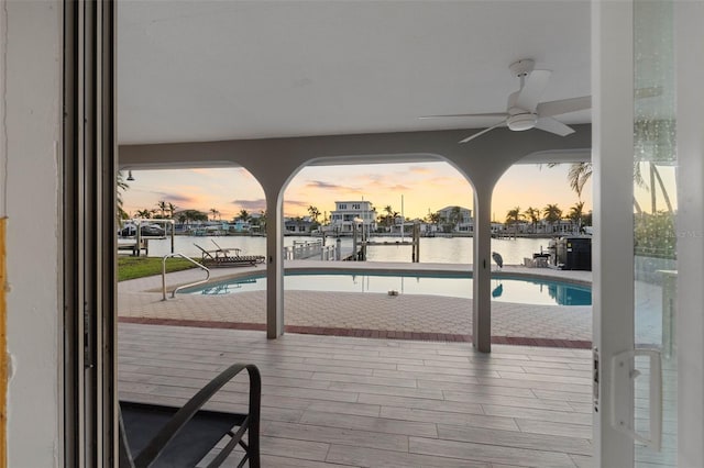 deck at dusk with ceiling fan, a water view, and a fenced in pool