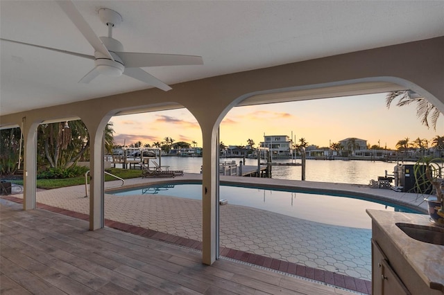 pool at dusk featuring a dock, ceiling fan, and a water view