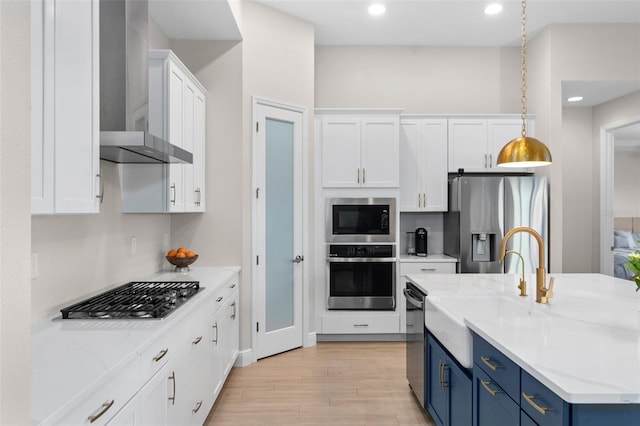 kitchen featuring white cabinets, wall chimney range hood, blue cabinetry, decorative light fixtures, and stainless steel appliances