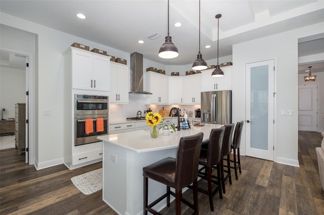 kitchen with appliances with stainless steel finishes, wall chimney exhaust hood, decorative light fixtures, a center island with sink, and white cabinetry