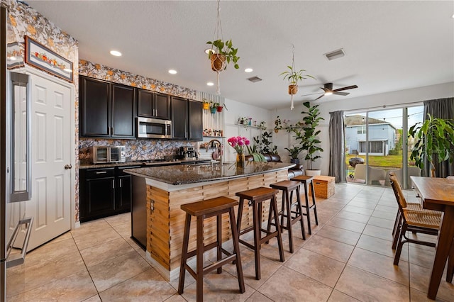 kitchen featuring sink, light tile patterned floors, an island with sink, dark stone counters, and decorative backsplash