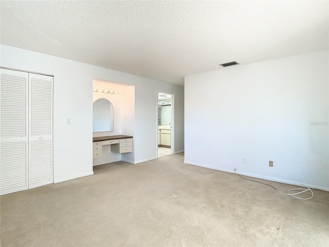 unfurnished bedroom with ensuite bath, a textured ceiling, light colored carpet, built in desk, and a closet