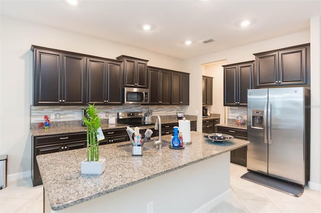 kitchen with appliances with stainless steel finishes, an island with sink, light stone countertops, and light tile patterned floors