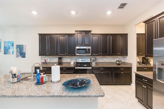 kitchen with light stone counters, stainless steel appliances, a center island with sink, light tile patterned floors, and decorative backsplash