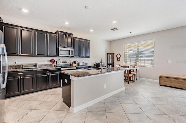 kitchen featuring dark stone counters, a center island with sink, light tile patterned floors, decorative light fixtures, and stainless steel appliances