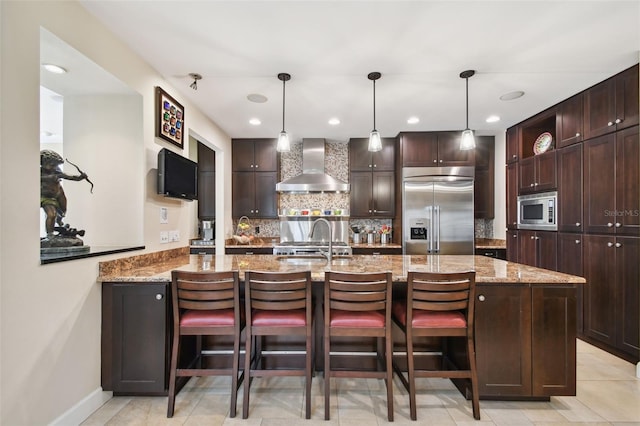 kitchen featuring wall chimney exhaust hood, a kitchen breakfast bar, built in appliances, and light stone counters