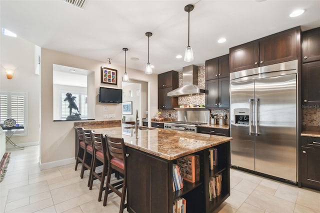 kitchen with an island with sink, built in fridge, wall chimney exhaust hood, light stone counters, and sink