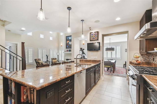 kitchen featuring stainless steel appliances, wall chimney range hood, decorative light fixtures, and a kitchen island with sink