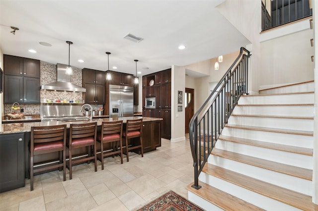 kitchen featuring decorative backsplash, a kitchen island with sink, hanging light fixtures, built in appliances, and wall chimney exhaust hood