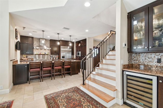 bar with dark brown cabinetry, beverage cooler, wall chimney exhaust hood, and built in appliances