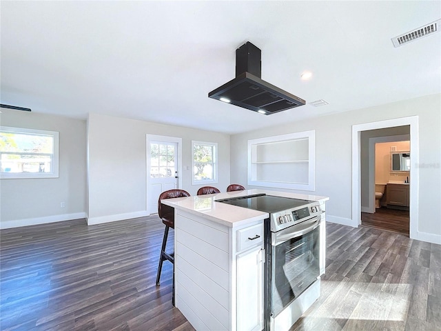 kitchen featuring stainless steel range with electric cooktop, a center island with sink, white cabinets, a breakfast bar area, and island exhaust hood