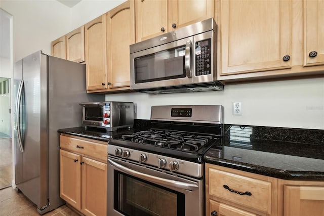 kitchen with light brown cabinetry, stainless steel appliances, light tile patterned floors, and dark stone countertops