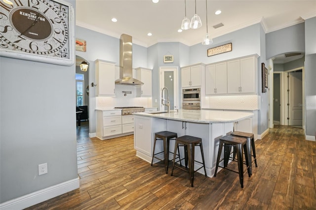 kitchen featuring wall chimney exhaust hood, white cabinetry, tasteful backsplash, sink, and a kitchen island with sink