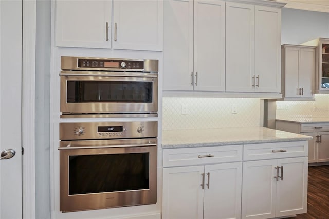 kitchen with dark hardwood / wood-style floors, stainless steel double oven, decorative backsplash, light stone countertops, and white cabinets