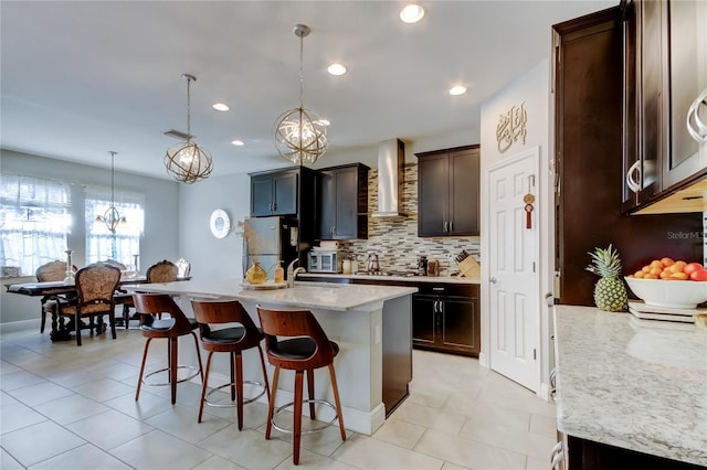 kitchen featuring wall chimney exhaust hood, light stone counters, decorative light fixtures, a center island with sink, and stainless steel refrigerator