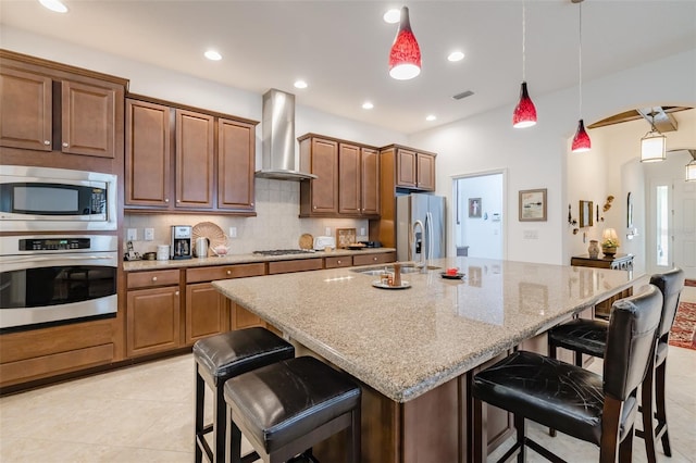 kitchen featuring decorative backsplash, a kitchen breakfast bar, wall chimney exhaust hood, stainless steel appliances, and a kitchen island with sink