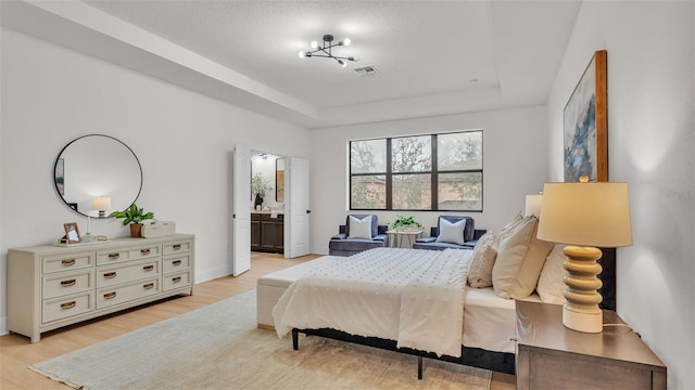 bedroom featuring a textured ceiling, connected bathroom, an inviting chandelier, light hardwood / wood-style floors, and a tray ceiling