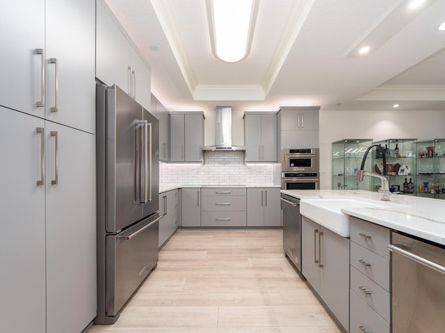 kitchen featuring appliances with stainless steel finishes, backsplash, a tray ceiling, wall chimney range hood, and gray cabinets