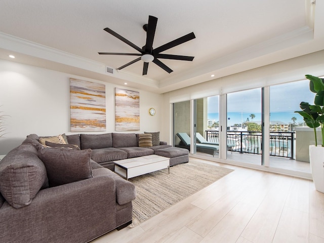 living room with ceiling fan, crown molding, a tray ceiling, a water view, and light wood-type flooring