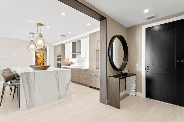 interior space featuring white cabinetry, wall chimney range hood, light stone counters, decorative light fixtures, and a kitchen bar