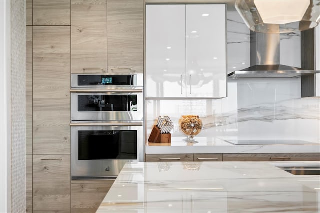 kitchen with light brown cabinets, wall chimney exhaust hood, light stone counters, double oven, and black electric cooktop