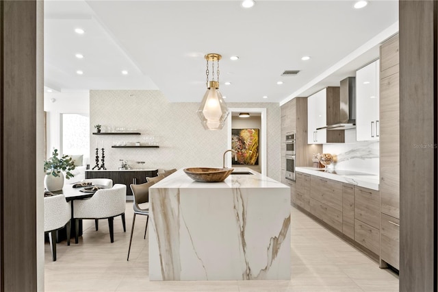 kitchen featuring sink, wall chimney exhaust hood, pendant lighting, a kitchen island with sink, and white cabinets