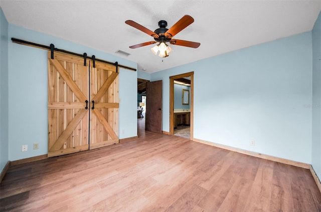 unfurnished bedroom with ensuite bathroom, ceiling fan, a barn door, and light wood-type flooring