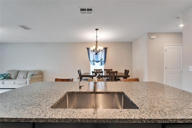 kitchen with sink, an inviting chandelier, light stone counters, an island with sink, and decorative light fixtures