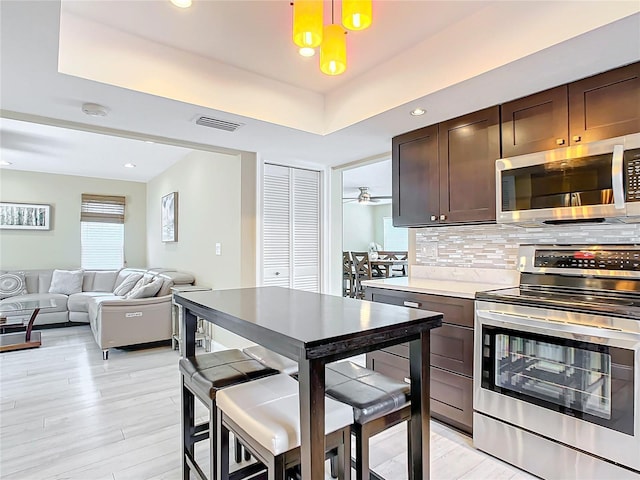 kitchen featuring stainless steel appliances, backsplash, a tray ceiling, dark brown cabinets, and light wood-type flooring