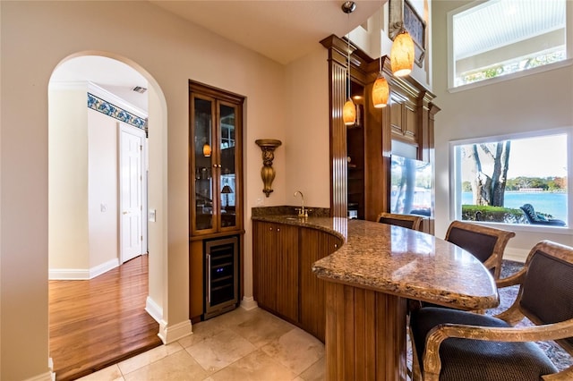 bar with sink, beverage cooler, light tile patterned floors, dark stone counters, and a water view