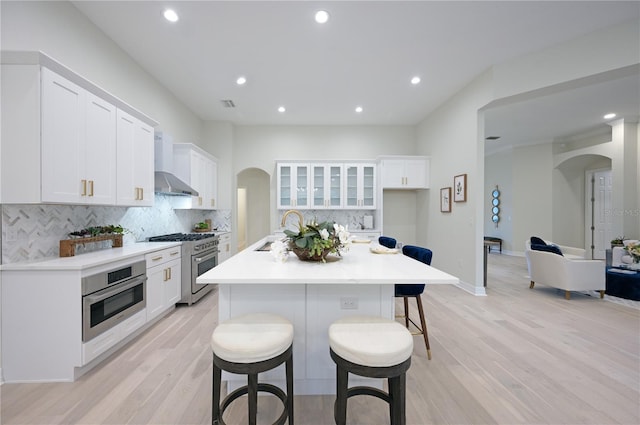 kitchen with wall chimney exhaust hood, stainless steel appliances, a kitchen breakfast bar, a center island with sink, and white cabinets