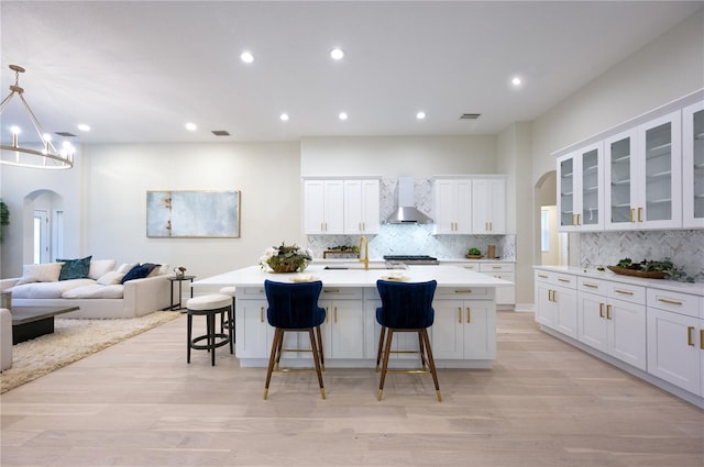 kitchen with a kitchen bar, wall chimney exhaust hood, a kitchen island with sink, pendant lighting, and white cabinetry