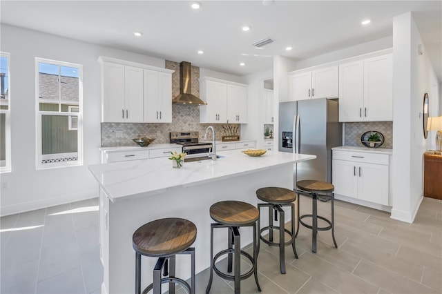 kitchen with white cabinets, wall chimney range hood, a kitchen island with sink, and stainless steel appliances