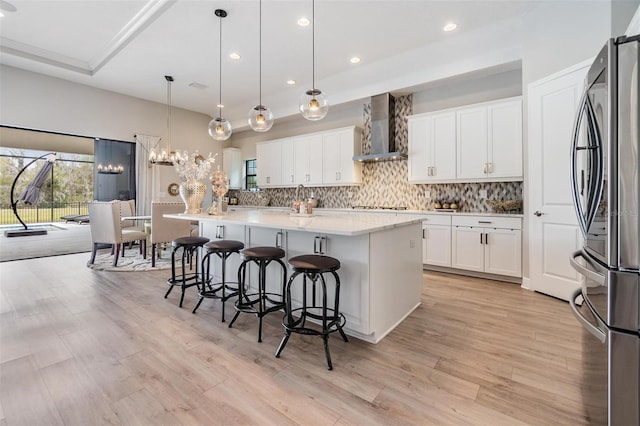 kitchen featuring a center island with sink, wall chimney exhaust hood, hanging light fixtures, and stainless steel refrigerator