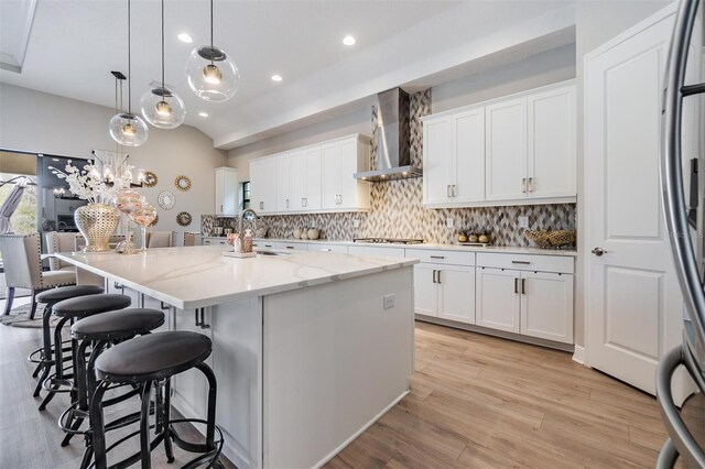 kitchen with white cabinetry, wall chimney exhaust hood, hanging light fixtures, light stone counters, and a center island with sink